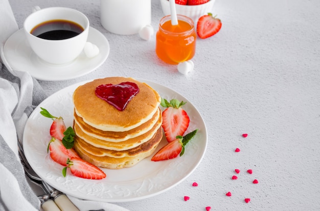 stapel pannenkoeken met een hart van jam bovenop met verse aardbeien en munt op een witte plaat op een lichte achtergrond.