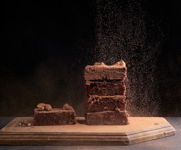 Stapel vierkant gebakken stukjes brownie besprenkeld met cacaopoeder, deeltjes bevroor in de lucht tegen een donker oppervlak