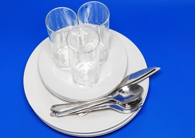 Stapel witte borden, glazen met vorken en lepels op zijden servet. Blauwe achtergrond