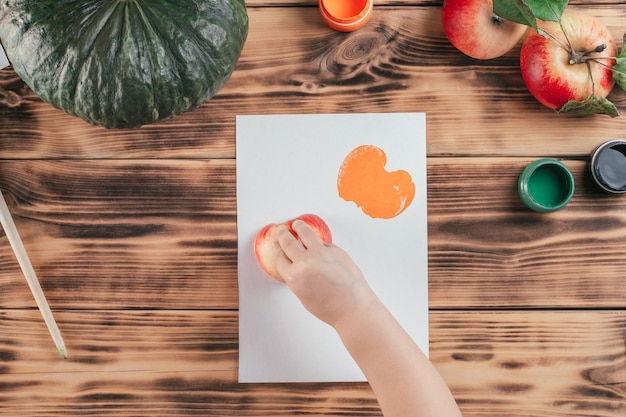 Stapsgewijze Halloween-tutorial voor kinderen met pompoen-appelprints. Stap 8: Kinderhand laat afdrukken op papier met de helft van de appel geschilderd in oranje gouacheverf. Bovenaanzicht