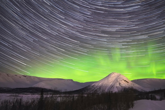 Star trails on night sky and green northern lights in snowy\
mountains at winter night.