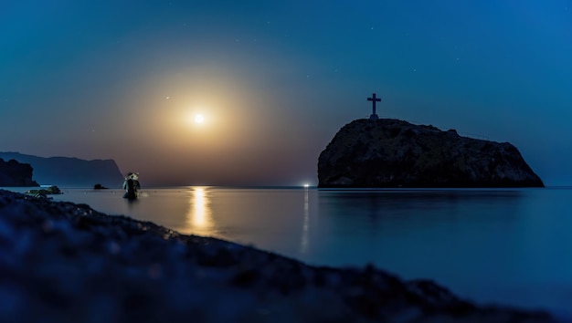 星空の夜、海の向こうに満月があり、正面に岩があります。