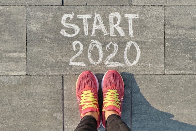 Start 2020, text on gray sidewalk with woman legs in sneakers, top view