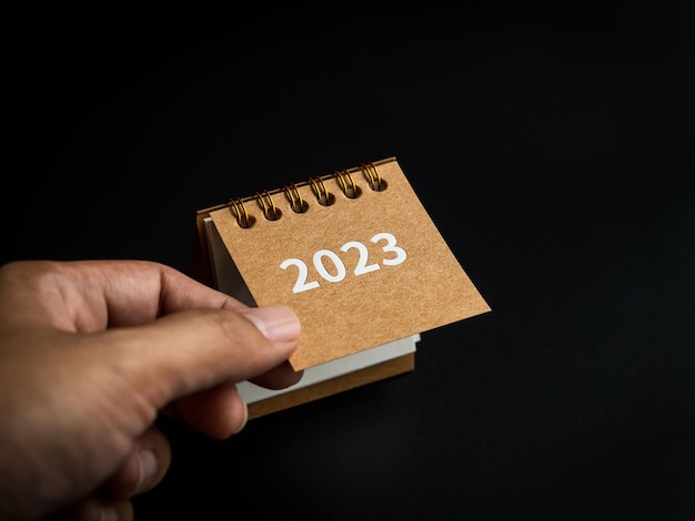 Start new goals happy new year 2023 background the 2023 year\
small beige desk calendar cover opens by hand to january the first\
month on black background minimal style