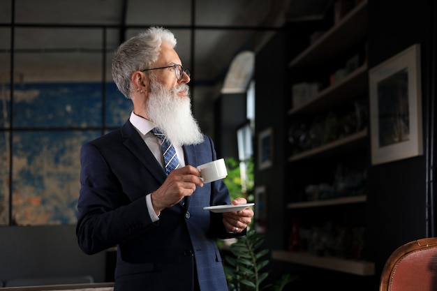 一杯のコーヒーで新しい仕事の日を始めましょう。彼の現代のオフィスに立っているコーヒーカップを保持しているハンサムな成熟した男。