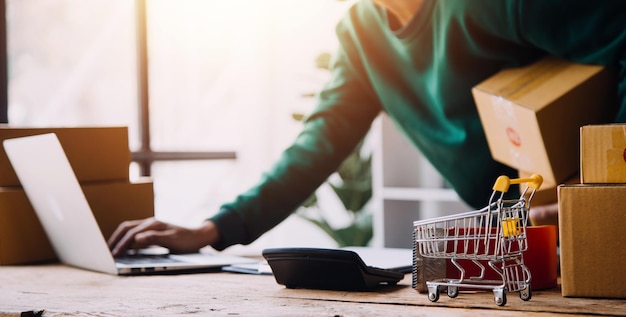 Startende mkb-ondernemer voor kleine bedrijven van freelance Aziatische vrouw met schort die laptop en doos gebruikt om online bestellingen te ontvangen en te beoordelen ter voorbereiding op het inpakken van verkoop aan klanten online mkb-bedrijfsideeën