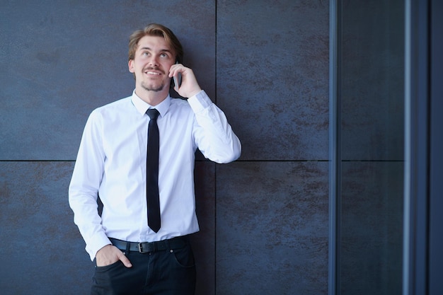 startende zakenman in een wit overhemd met een zwarte stropdas die mobiele telefoon gebruikt terwijl hij voor een grijze muur staat tijdens een pauze van het werk buiten
