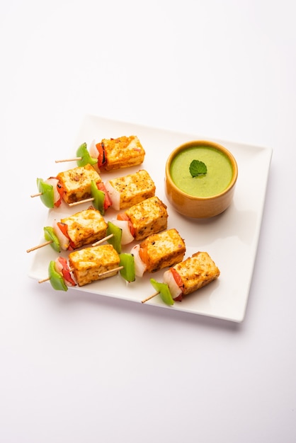 Starter snack Paneer Tikka with stick in plate with green chutney isolated on white. Indian cuisine dish with grilled cottage cheese with vegetables and spices