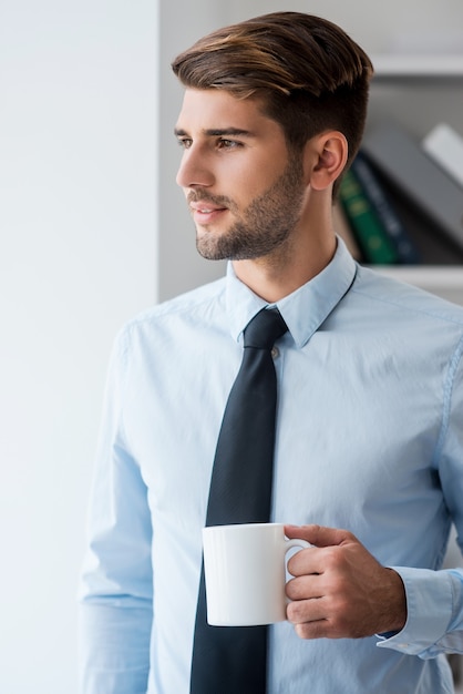 淹れたてのコーヒーで一日を始めましょう。コーヒーカップを保持しているシャツとネクタイの思いやりのある若い男