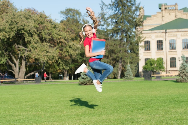 Starting school in good mood Happy schoolgirl jump to music on green grass Active little child in headphones hold school books Back to school School holidays Summer vacation Primary education