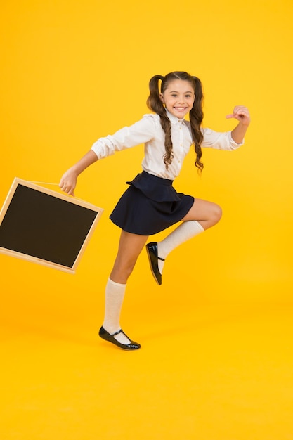 笑顔で学校を始める黄色の背景で学校に行く幸せな小さな子供学校の制服を着たアクティブな女の子が黒板を持って学校のシーズンに戻るコピースペース