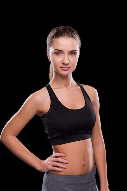 Starting training. Beautiful young woman holding hand on hip and looking at camera while standing against black background