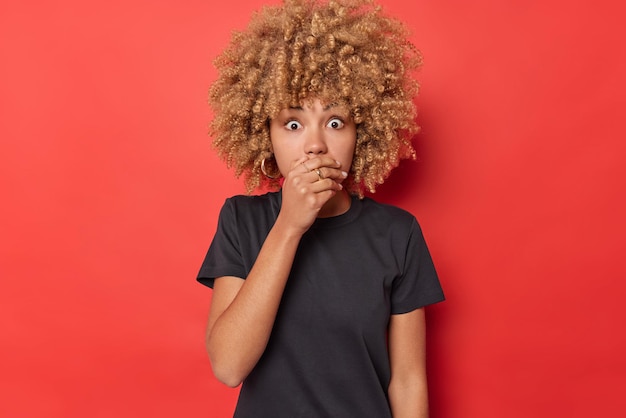 Startled frightened curly woman covers mouth with hand stares at something horrible tries to be speechless wears casual black t shirt isolated over vivid red background. Omg dont drop any word