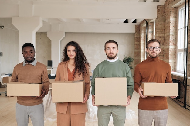 Startup company employees standing with cardboard boxes in row and looking at camera they moving to new office