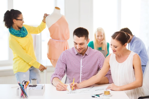 startup, education, fashion and office concept - smiling designers drawing sketches and adjusting dress on mannequin in office