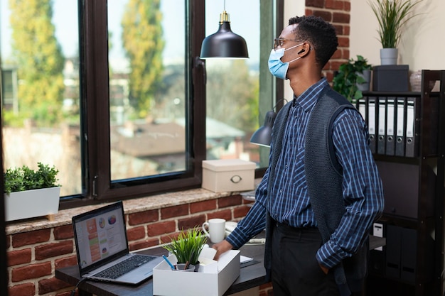 Startup employee wearing covid mask looking out the window with regret after being fired from work. Laid off marketing specialist standing next to desk with personal belongings and laptop.