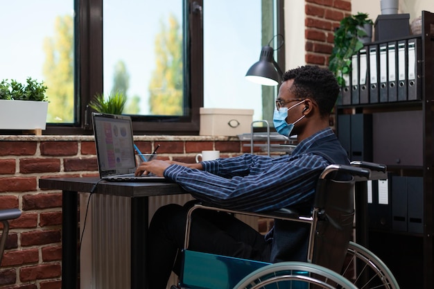 Photo startup employee who uses wheelchair wearing covid face mask working on laptop with business analytics. african american marketing specialist analyzing key performance indicators.