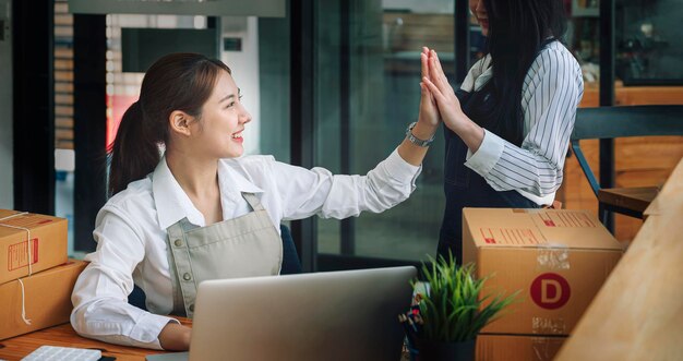 Startup small busines SME. Beautiful asian entrepreneur giving hi five bump hand with her friend and using laptop computer check online order. Successful SME business online concept.