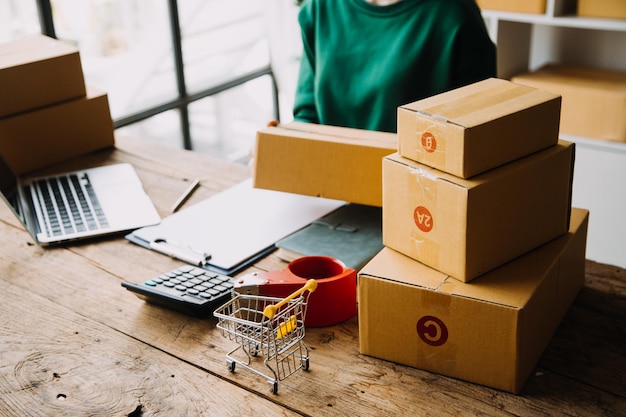 Стартап-предприниматель малого бизнеса или внештатная азиатка, использующая ноутбук с коробкой Молодая успешная азиатка с рукой поднимает упаковочную коробку онлайн-маркетинга и концепцию доставки МСП