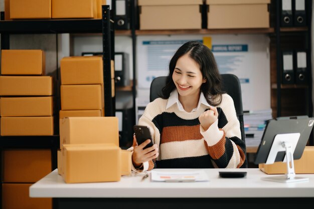 Startup small business sme entrepreneur owner asian woman using smartphone or tablet taking receive and checking online purchase shopping order to preparing pack product box