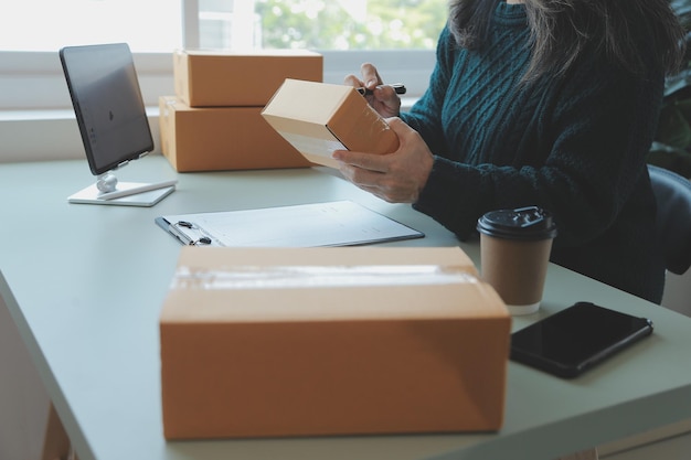 Photo startup sme small business entrepreneur of freelance asian woman wearing apron using laptop and box to receive and review orders online to prepare to pack sell to customers online sme business ideas