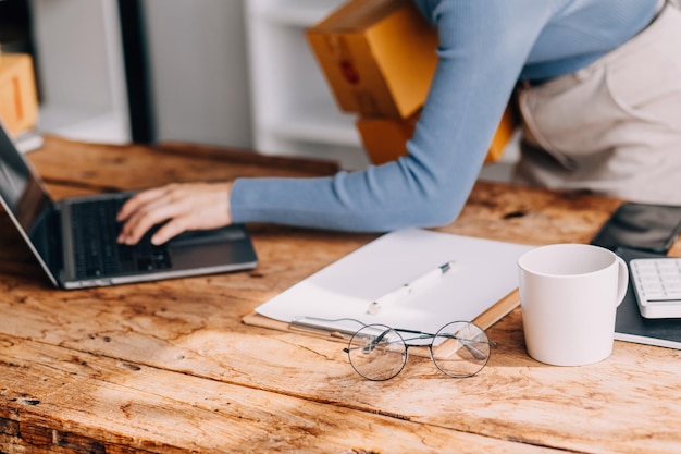 Photo startup sme small business entrepreneur of freelance asian woman wearing apron using laptop and box to receive and review orders online to prepare to pack sell to customers online sme business ideas