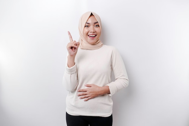 Starving Asian Muslim woman wearing a headscarf pointing up at the copy space above her isolated by a white background