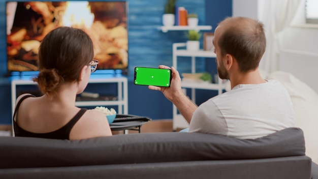 Static tripod shot of couple enjoying social media videos on smartphone with green screen in modern living room. Man holding smartphone with chroma key watching online video content with girlfriend.