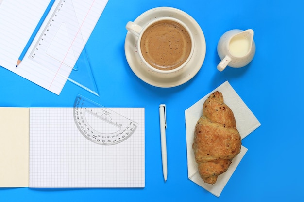 Foto articoli di cancelleria e caffè con cornetto su sfondo blu quaderni con fogli bianchi