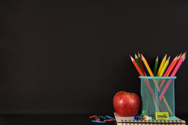 Stationery or school supplies with books, color pencils, clips and red apple on black background.