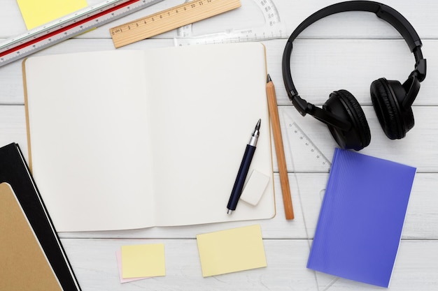 Stationery supplies in modern office workspace. Top view on open blank notebook, pen, pencil, rulers, small notepads and memo pads, headphones on white wooden desktop, flat lay, copy space, mockup