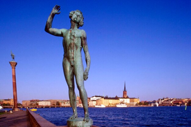 Statue by river with riddarholm church in background against blue sky