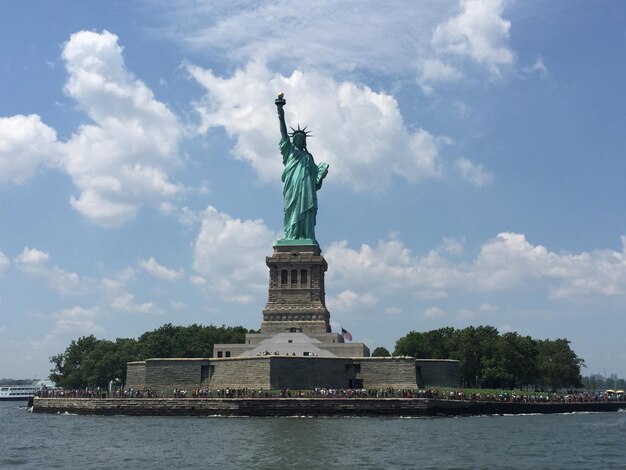 Photo statue of liberty by river against cloudy sky
