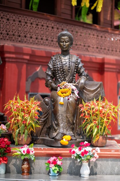 A statue of a woman sits in front of a red building with flowers around it.