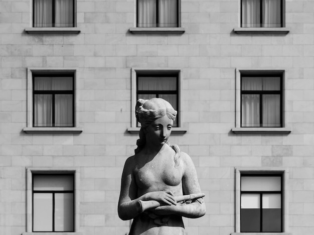 A statue of a woman with a fish in her hands stands in front of a building with windows.