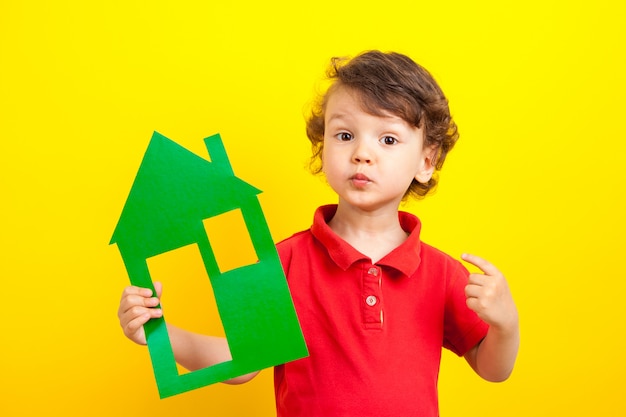 Stay home. cheerful cute baby boy holds a mock-up of a cardboard green house and points a finger at it. Laughs and smiles. On a studio yellow , isolated. Positive at home.