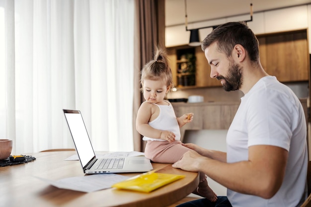 A stay at home dad is working on a laptop while his toddler is helping him