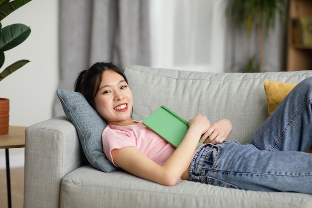 Оставайся дома, хобби Веселая азиатка, лежащая на диване с книгой, смотрящая и улыбающаяся в пространство для копирования камеры