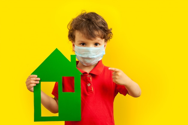 Stay home. A masked boy holds a green house model in his hands and points a finger at it.