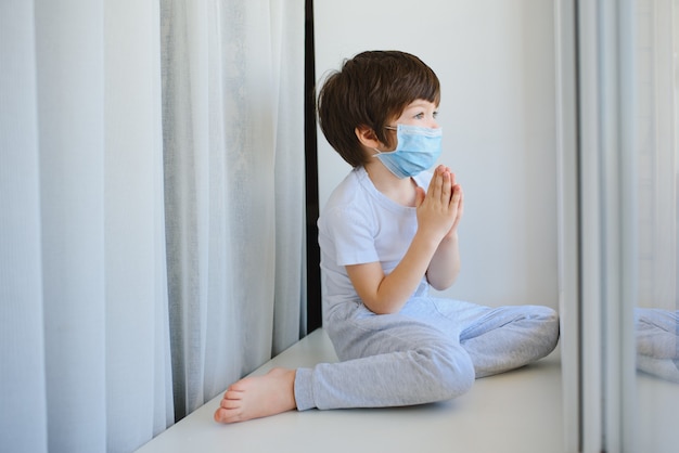 코로나 바이러스 전염병 예방을 위해 집에서 격리하십시오. 보호용 의료 마스크를 쓴 어린이와 그의 테디 베어는 창턱에 앉아 창 밖을 내다 봅니다.