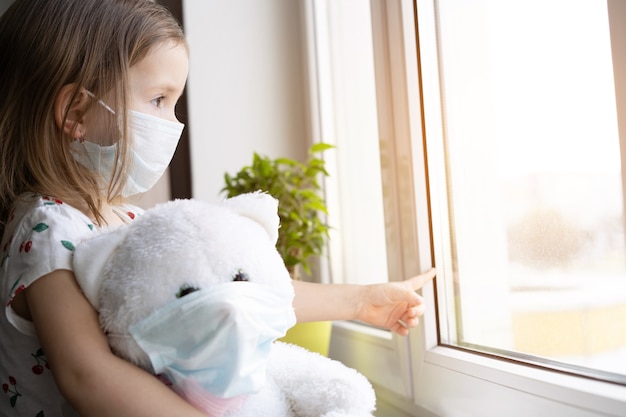 Stay at home quarantine coronavirus pandemic prevention. Sad child and his teddy bear both in protective medical masks sits on windowsill and looks out window. COVID-2019 disease concept