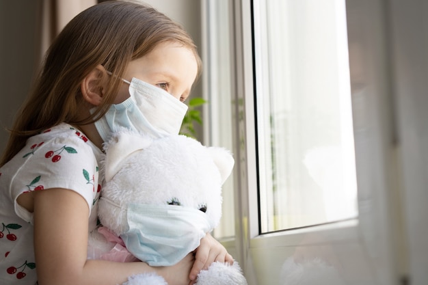 Stay at home quarantine coronavirus pandemic prevention. Sad child and his teddy bear both in protective medical masks sits on windowsill and looks out window. COVID-2019 disease concept