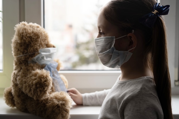 Stay at home quarantine coronavirus pandemic prevention. Sad child and his teddy bear both in protective medical masks sits on windowsill and looks out window. COVID-2019 disease concept