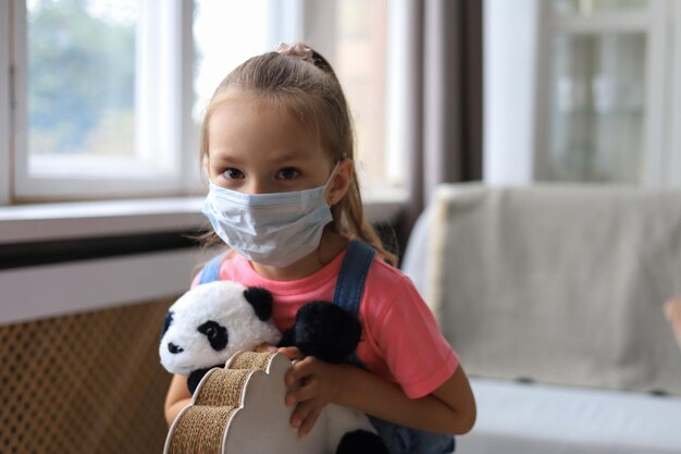 Stay at home quarantine coronavirus pandemic prevention Sad child in protective medical mask and her panda bear sit on rocking horse looking at camera