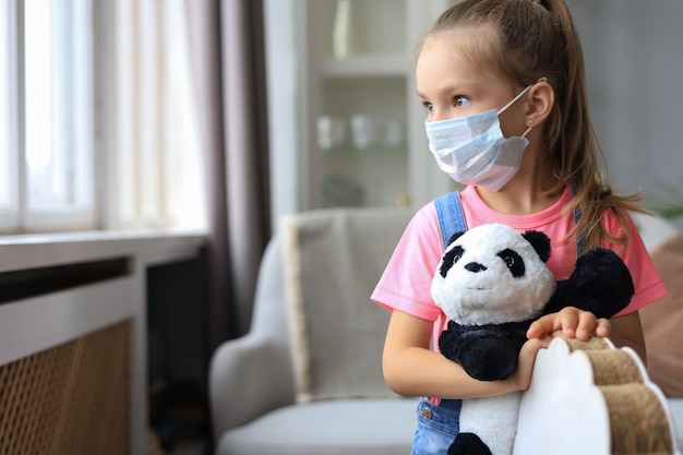 Resta a casa quarantena prevenzione della pandemia di coronavirus il bambino triste in maschera medica protettiva e il suo orso panda si siedono su un cavallo a dondolo guardando fuori dalla finestra