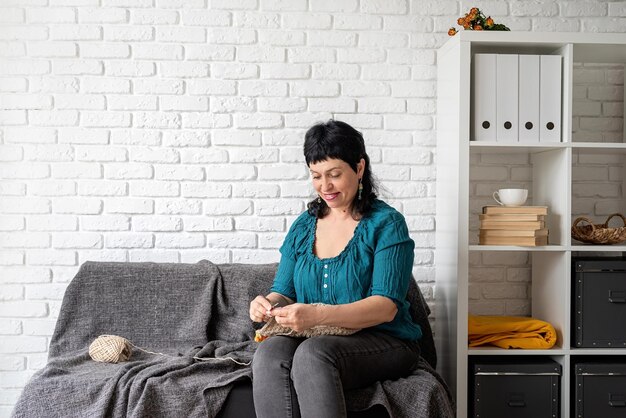 Stay home smiling middle aged woman enjoying being at home and knitting sitting on the sofa