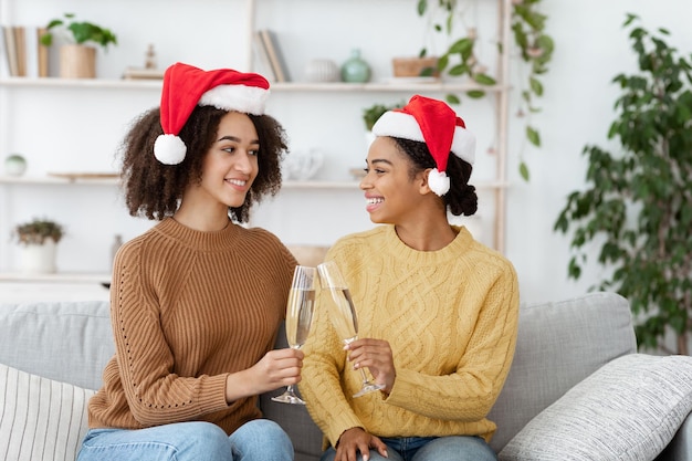 Оставайтесь дома с семьей или друзьями, чтобы отпраздновать Рождество и Новый год. Счастливые афро-американские молодые женщины в свитерах и шляпах Санта-Клауса чокаются с шампанским в интерьере уютной гостиной