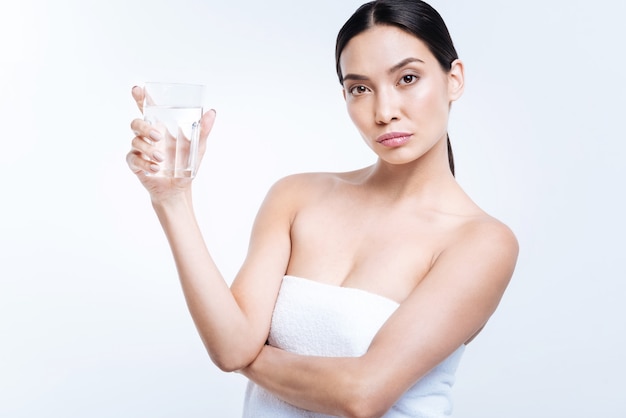 Stay hydrated. Charming young woman showing a glass of water while standing wrapped in a towel against a white wall