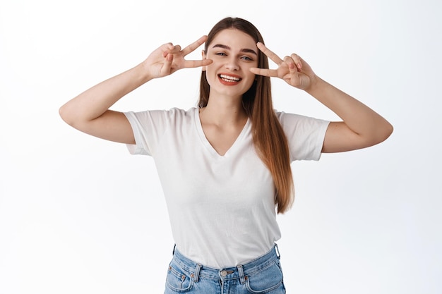 Stay positive. Smiling beautiful girl shows peace v-sign and looks happy at camera, express bright joyful emotions, enjoy spring, standing in t-shirt over white background
