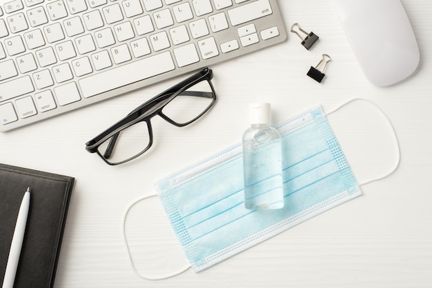 Stay safe at work concept. Top above overhead flat lay photo of office workplace with surgical filter mask and antiseptic laying on wooden white desk